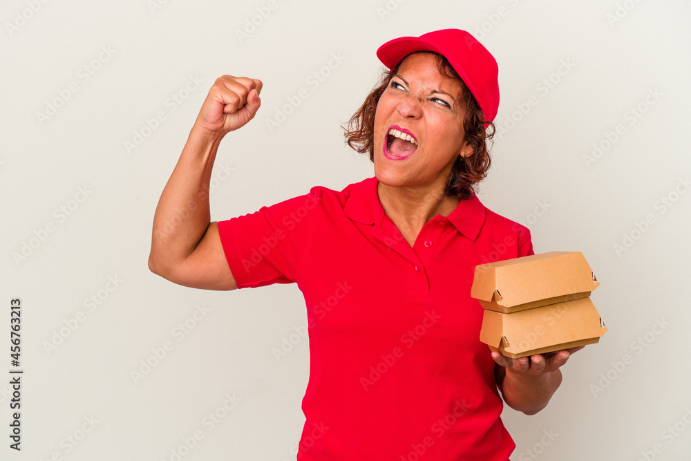 Middle age delivery woman taking burguers isolated on white background raising fist after a victory, winner concept.