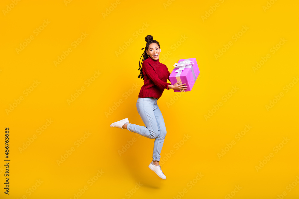 Full length high of shiny adorable dark skin woman dressed red sweater jumping holding gift box isolated yellow color background