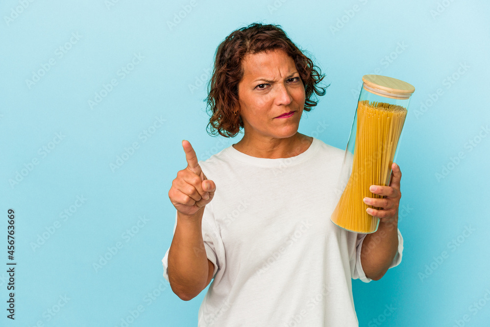 Middle age latin woman holding pasta jar isolated on blue background showing number one with finger. Middle age latin woman holding pasta jar isolated on blue background showing number one with finger.