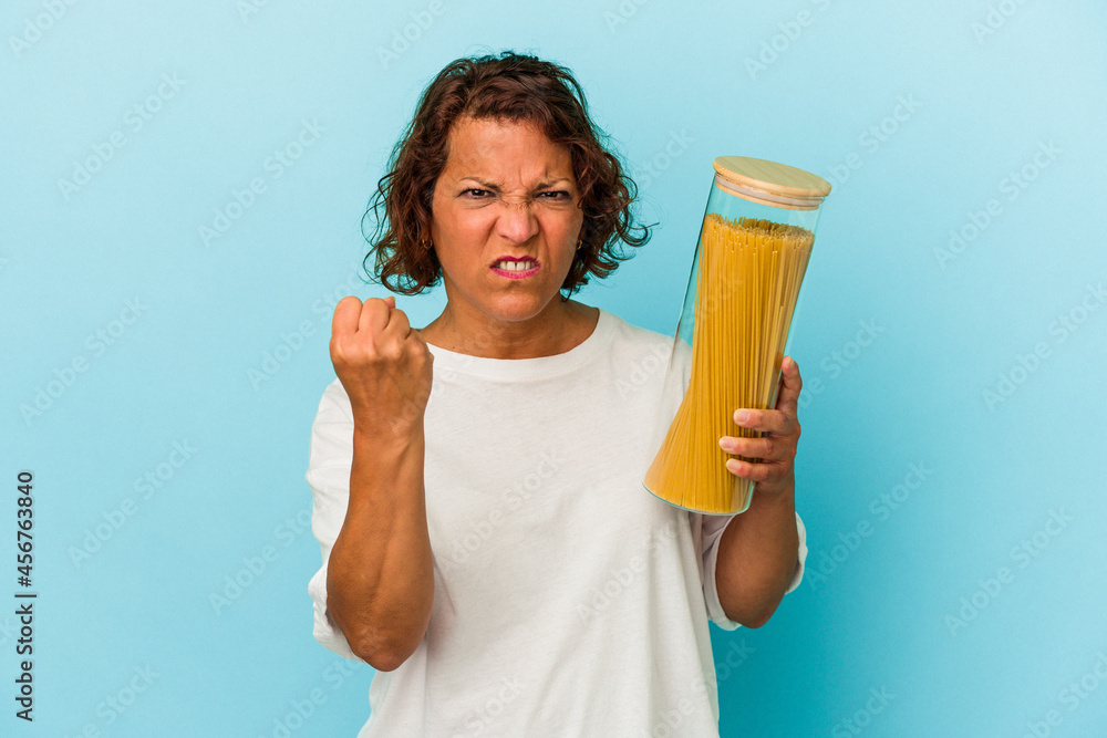 Middle age latin woman holding pasta jar isolated on blue background showing fist to camera, aggressive facial expression. Middle age latin woman holding pasta jar isolated on blue background showing fist to camera, aggressive facial expression.