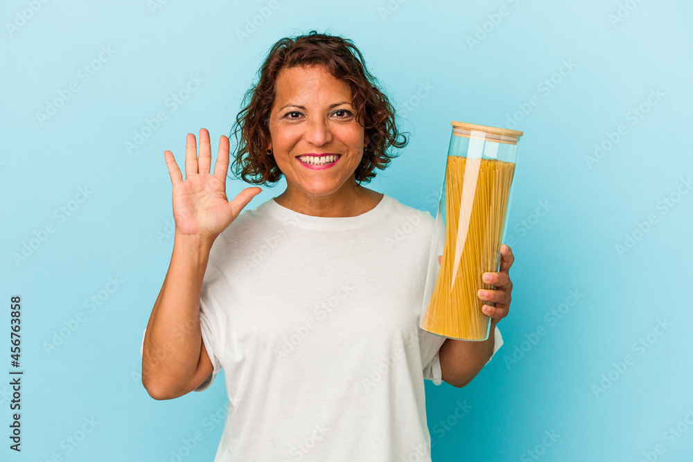 Middle age latin woman holding pasta jar isolated on blue background smiling cheerful showing number five with fingers. Middle age latin woman holding pasta jar isolated on blue background smiling cheerful showing number five with fingers.