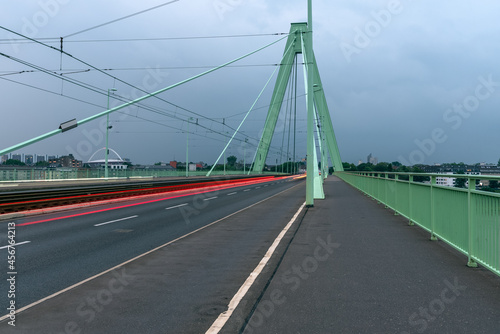 Fototapeta Naklejka Na Ścianę i Meble -  Empty walkway and bicycle lane along a road over a suspension bridge on a cloudy summer evening. A tramway runs in the middle of the road. Light trails left by passing cars..