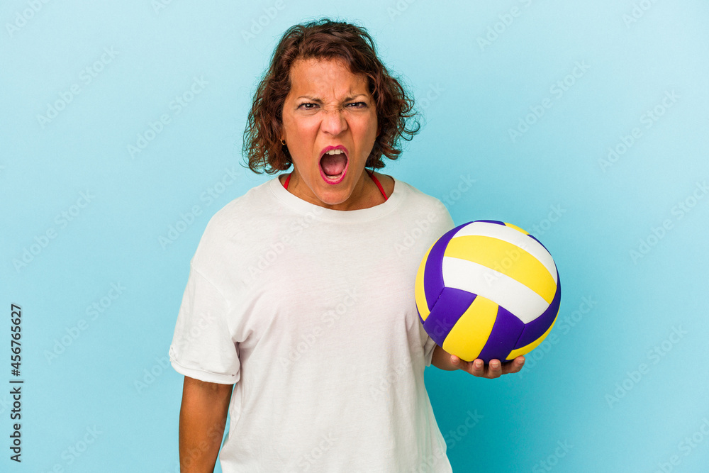 Middle age latin woman playing volleyball isolated on blue background screaming very angry and aggressive.
