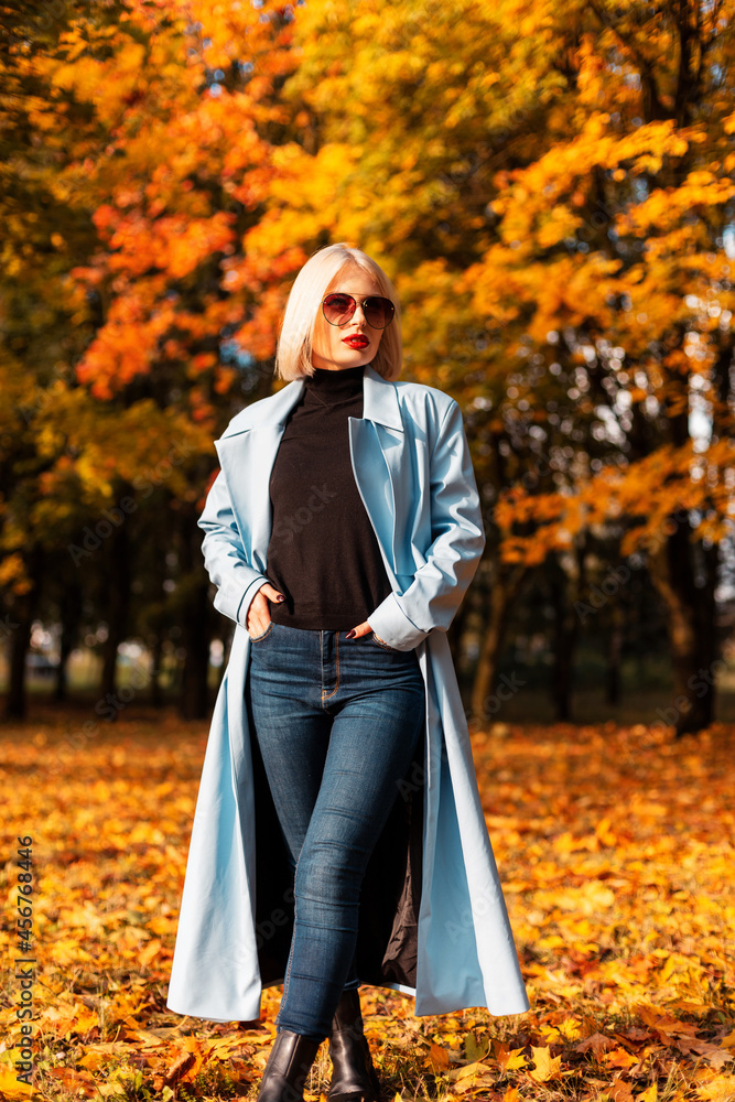 Stylish beautiful young woman in a fashion blue coat with jeans and sweater walks in an autumn yellow park with fall foliage on a sunny day