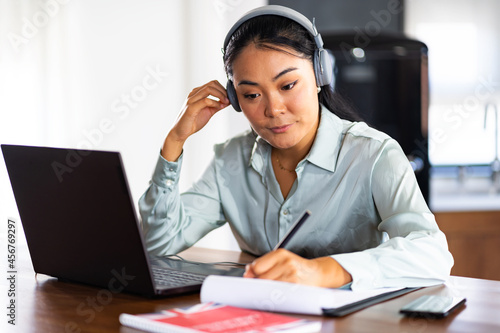 Young Asian woman attending online foreign language classes. Sitting in front of laptop computer with headphones listening course and taking notes.
