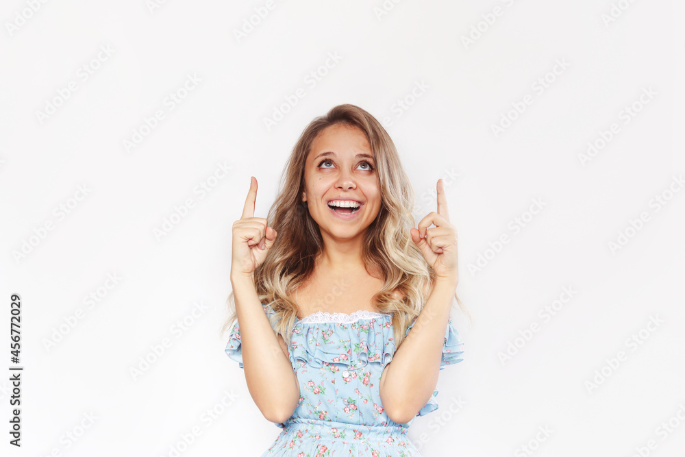 A young caucasian excited smiling blonde woman with wavy hair in a blue dress points up to a empty copy space for text or design with two fingers presenting product isolated on a white background A young caucasian excited smiling blonde woman with wavy hair in a blue dress points up to a empty copy space for text or design with two fingers presenting product isolated on a white background