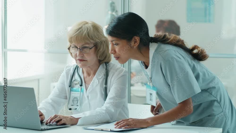 Tilt down shot of young Hispanic nurse and senior Caucasian female doctor discussing something on laptop while working together in clinic Tilt down shot of young Hispanic nurse and senior Caucasian female doctor discussing something on laptop while working together in clinic