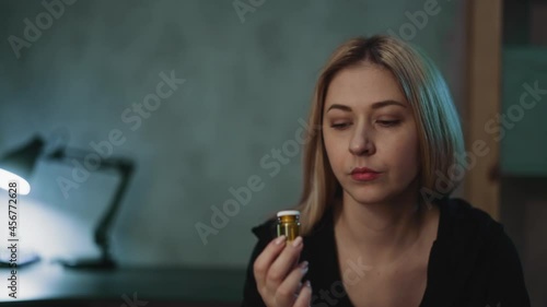 Thoughtful woman turns glass drug bottle with unregistered nutritional products doubting in doctor prescription in semi-dark room closeup slow motion photo