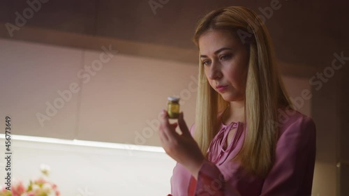 Thoughtful lady in pink blouse looks at cyanide powder in bottle planning murder or suicide in kitchen at home close view slow motion photo