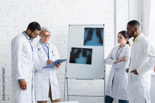 middle aged asian doctor pointing at clipboard near lungs fluorography and multiethnic colleagues photo