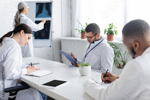 mature doctor near flip chart with lungs x-rays and multiethnic colleagues writing in meeting room