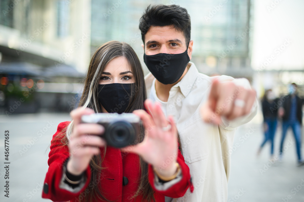 Happy tourists with covid or coronavirus masks couple whalking together in a city and photographing in the direction of the camera