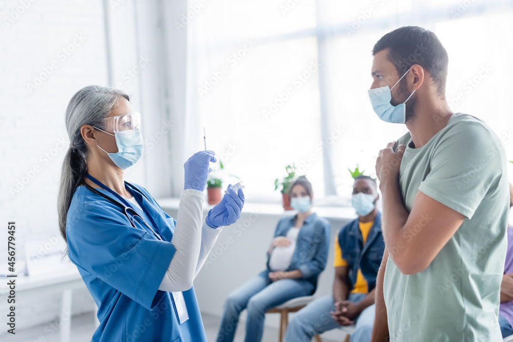 Fototapeta premium mature asian nurse in medical mask holding vaccine near young man and blurred multiethnic patients