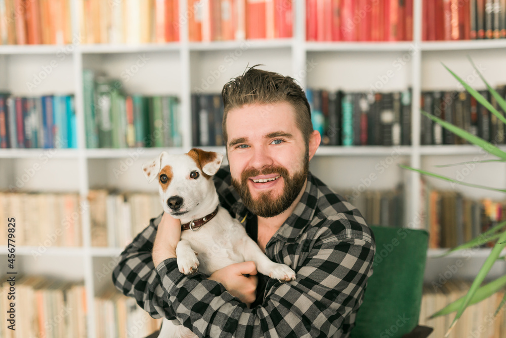 Happy male owner of jack russell terrier dog, feels responsibility of caring about pet, standing against bookshelves background. People and relationship with animals