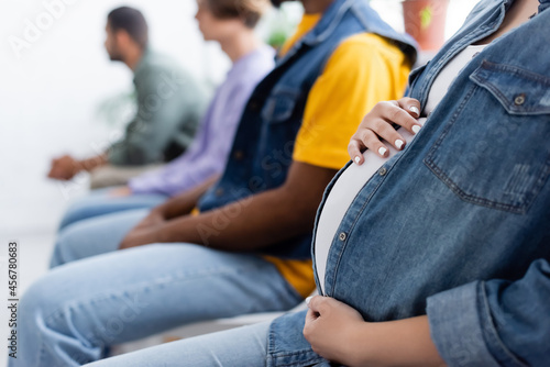 cropped view of pregnant woman touching belly while sitting in line with blurred multiethnic men, vaccination concept