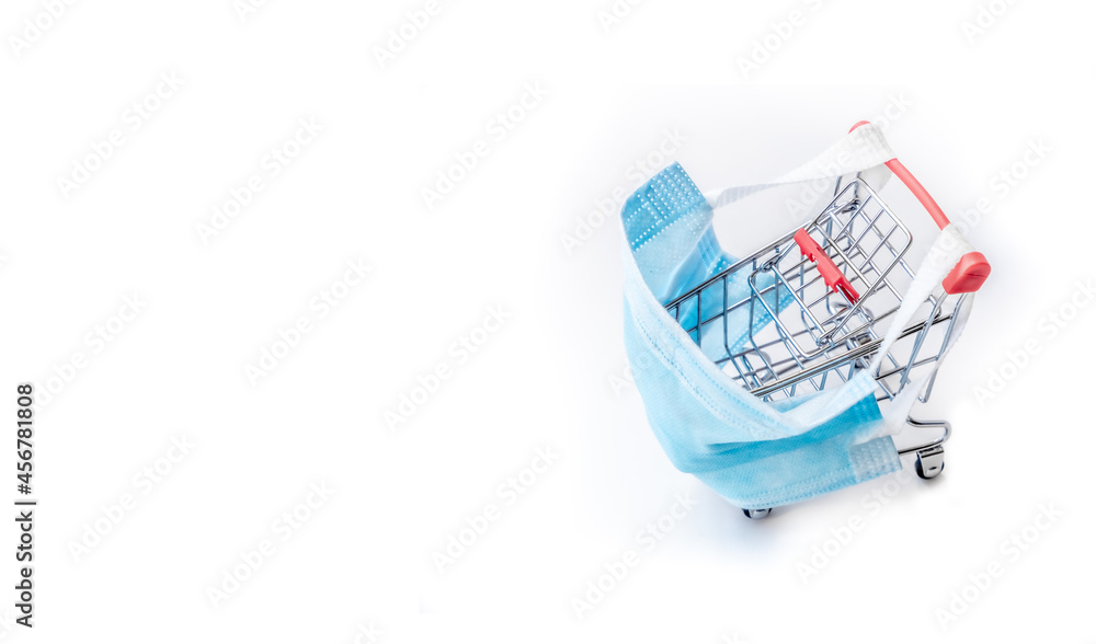 Shopping cart with blue protective medical mask against coronavirus. Safe and online shopping on quarantine concept. Light background, copy space, isolate. Shopping cart with blue protective medical mask against coronavirus. Safe and online shopping on quarantine concept. Light background, copy space, isolate.