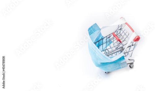 Shopping cart with blue protective medical mask against coronavirus. Safe and online shopping on quarantine concept. Light background, copy space, isolate.