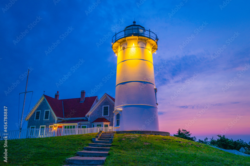Glowing golden light of Nobska Lighthouse illuminating the green hill before sunrise. Pink and blue sky at dawn over the Atlantic Ocean.