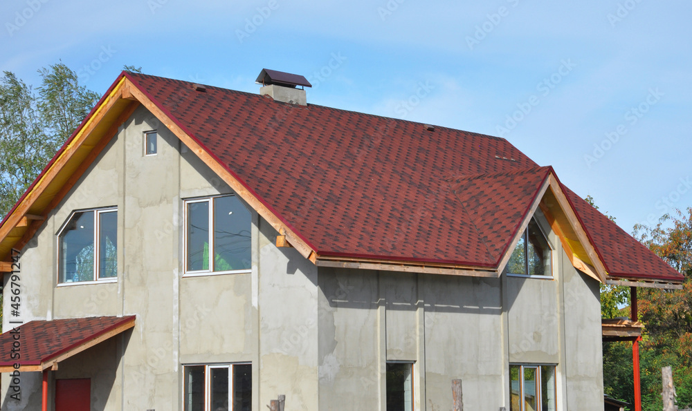 A house under construction with a red asphalt shingled roof, a chimney, attic windows, incomplete soffit and fascia board, and stucco, plastered walls. A house under construction with a red asphalt shingled roof, a chimney, attic windows, incomplete soffit and fascia board, and stucco, plastered walls.