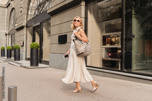 Beautiful blonde caucasian mature woman walking while shopping on the city street, carrying handbag outdoors. Middle-aged businesswoman buying goods clothes