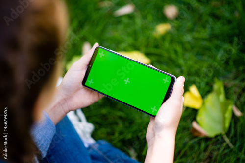 A young girl in blue jeans sits on the grass with yellow leaves and looks at the phone. Phone with green screen.