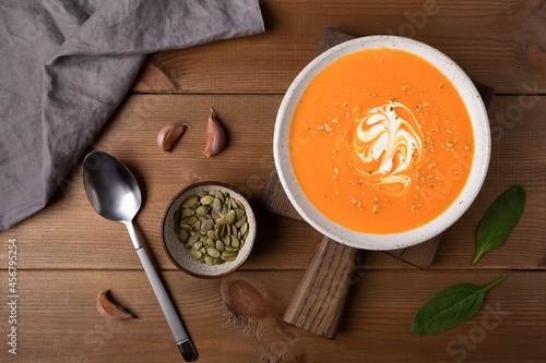 Homemade pumpkin soup in a white plate with cream, spoon, pumpkin seeds, garlic, napkin flat lay on brown wooden rustic background.