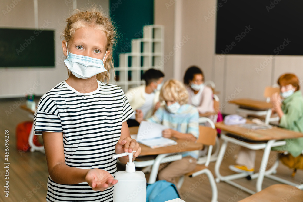 Small kid schoolgirl pupil student in protective medical face mask using sanitizer to protect from Covid19 coronavirus in class lesson at school. Lockdown restrictions.