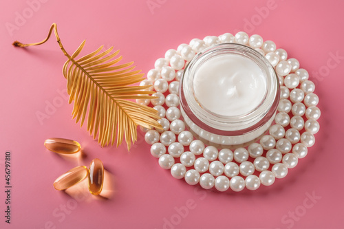 Face cream in an open glass jar and white pearl beads around, few essential oil capsules and golden yellow dry leaf on a pastel pink background. Beauty cream and dry skin care cosmetics.
