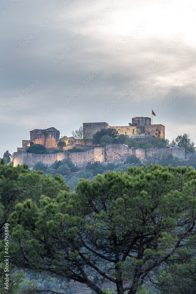 Old stone castle with flag of catalonia on top of a mountain at dusk illuminated with artificial light and sky with gray clouds.