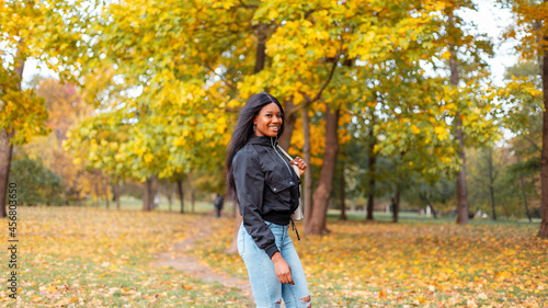 Smiling young black girl in fashion casual clothes with stylish jacket and jeans walking in an autumn park with yellow fall foliage