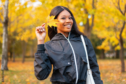 Young happy beautiful black girl with a cute smile in fashionable casual clothes holding a yellow autumn leaf is walking in the park with bright colorful fall foliage
