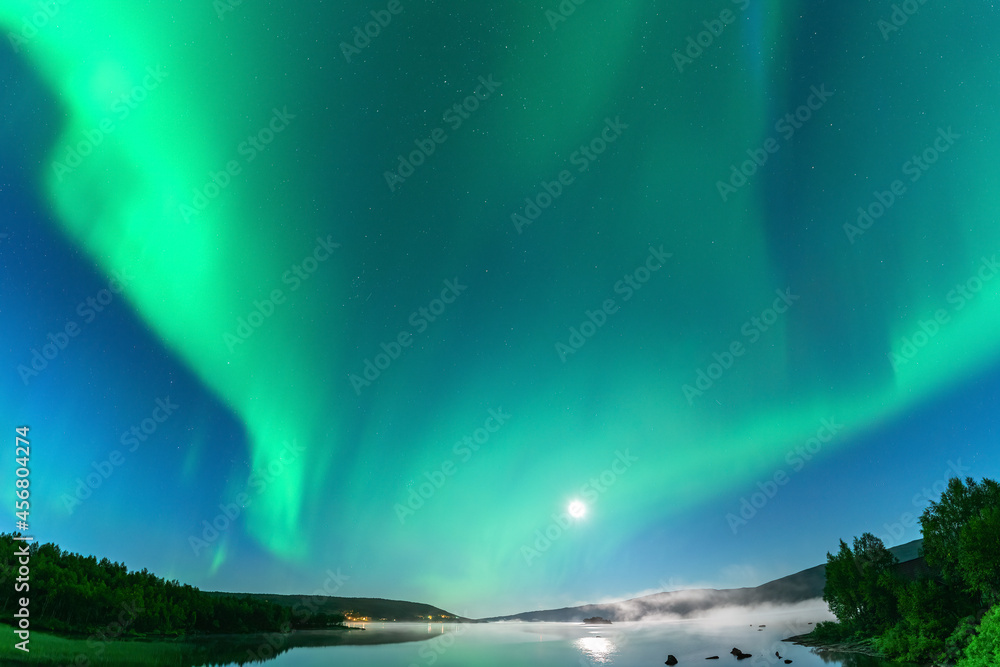 Panoramic Aurora borealis, Northern green lights with full moon and stars in the night sky over mountain lake, mirrored reflection in water, night mist. Night road, Joesjo, Lappland, Northern Sweden
