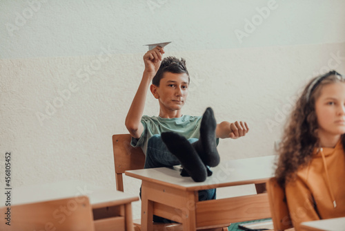 A naughty boy with his feet on a desk sits in the classroom and throws a plane. Selective focus photo
