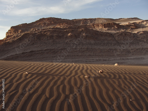 Sand drawn by the wind, with some little rocks showing long shadows caused by afternoon light and a stonewall behind it, in Valle de la Luna, Atacama Desert, Chile.
