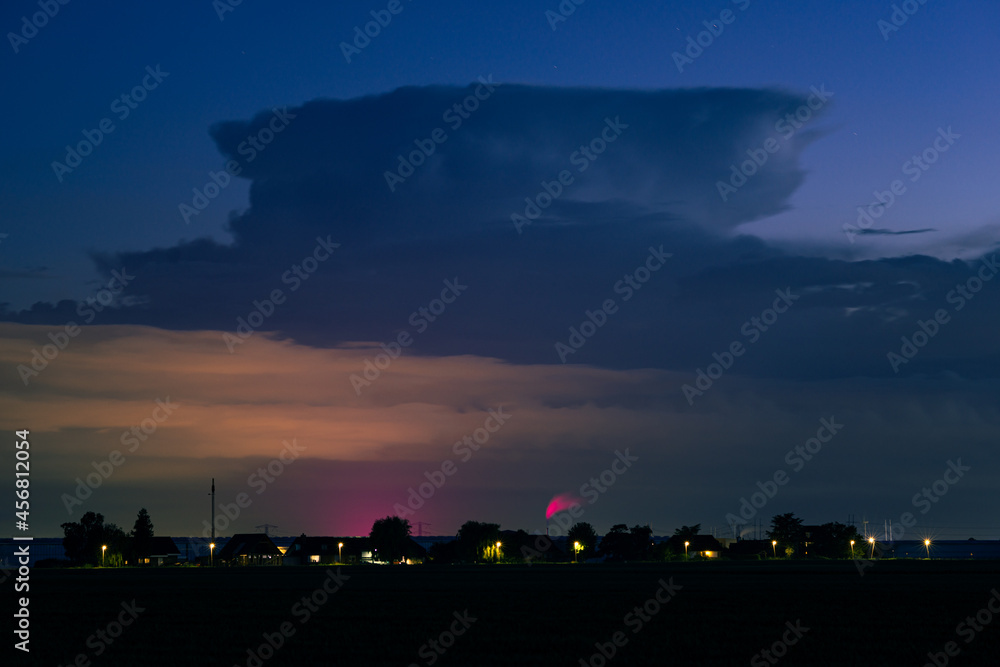 Storm cloud in the distance. Lower and nearby clouds are illuminated in orange and purple by the light from greenhouses. Storm cloud in the distance. Lower and nearby clouds are illuminated in orange and purple by the light from greenhouses.