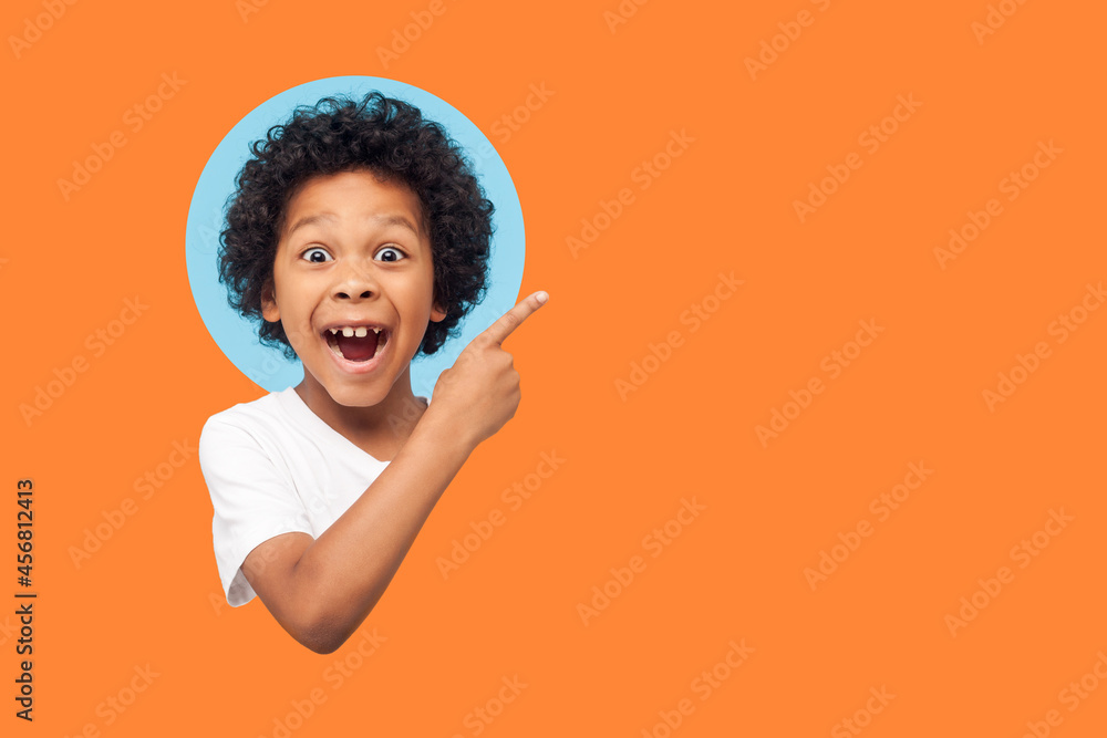 © khosrork - Wow look advertise here! Portrait of amazed little boy with curly hair pointing to empty place on background showing copy space for promotional ad. indoor isolated in a round hole on orange background © khosrork - Wow look advertise here! Portrait of amazed little boy with curly hair pointing to empty place on background showing copy space for promotional ad. indoor isolated in a round hole on orange background