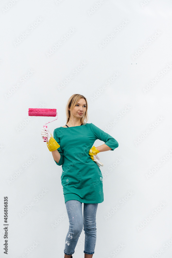 A young girl poses with a pink roller before painting a white wall. Repair of the interior. A young girl poses with a pink roller before painting a white wall. Repair of the interior.