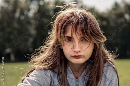 Casual portrait of teenager girl in a park, Selective focus. The model is with light make up and fake freckles in grey clothes. Looking at the viewer. Dark green trees in the background