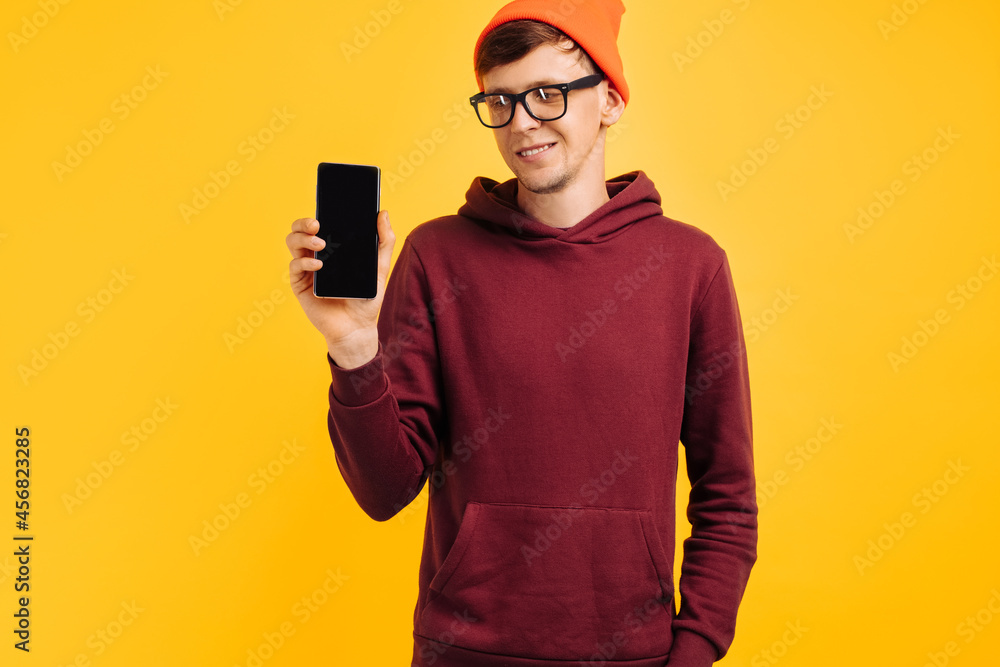 cheerful young guy in an orange hat with glasses and a red sweater shows phone screen close-up, on a yellow background cheerful young guy in an orange hat with glasses and a red sweater shows phone screen close-up, on a yellow background