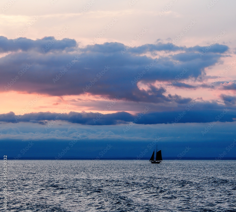 Fototapeta premium Sailboat with two sails (sloop?) in silhouette at horizon against dramatic, dark blue and dusky orange sunset and lightly choppy waters. 