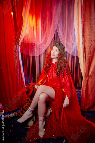 Young woman during a stylized theatrical circus photo shoot in a beautiful red location. Girl posing on stage with curtain