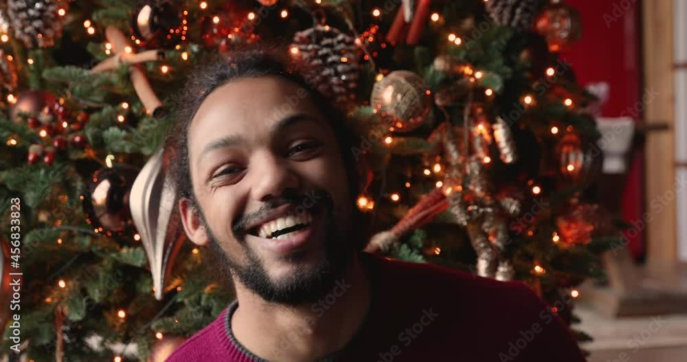 Head shot close up young handsome african american man holding video web camera call with festive decorated Christmas tree on background, congratulating family or friends with New Year distantly. Head shot close up young handsome african american man holding video web camera call with festive decorated Christmas tree on background, congratulating family or friends with New Year distantly.