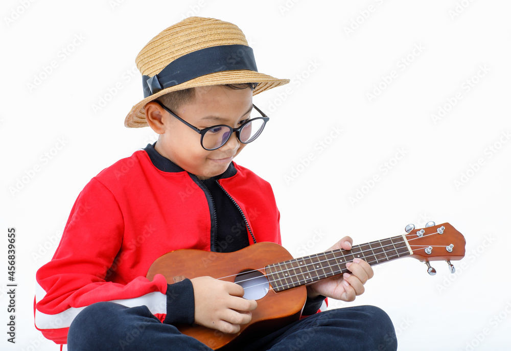 Lovey cutout portrait of young Asian boy wearing brown hat, glasses, red shirt, and long black pants happily sitting with smile while enjoy performing as musician by playing small guitar Lovey cutout portrait of young Asian boy wearing brown hat, glasses, red shirt, and long black pants happily sitting with smile while enjoy performing as musician by playing small guitar