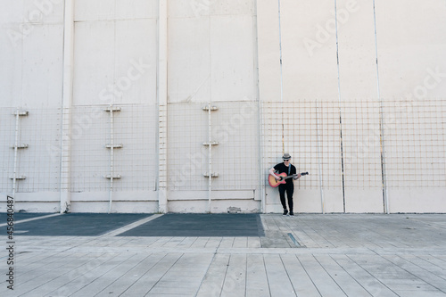 Distant shot of an albino man leaning against a wall while playing a song on his guitar. Concept of loneliness, remoteness, abandonment.