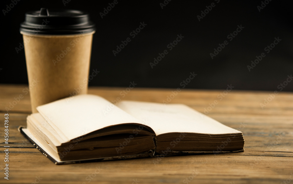 Open book and a cup of coffee in a disposable paper cup on a wooden table Open book and a cup of coffee in a disposable paper cup on a wooden table
