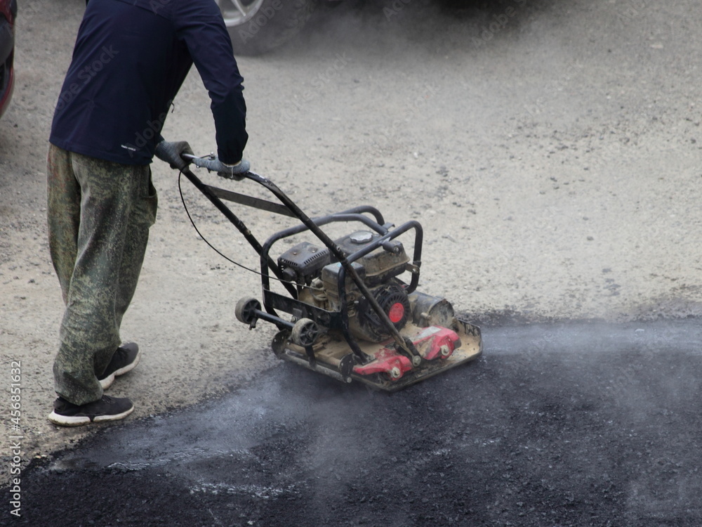 Obraz premium A worker rams new asphalt with a manual motor asphalt rammer, asphalting by the pit method, local road repair