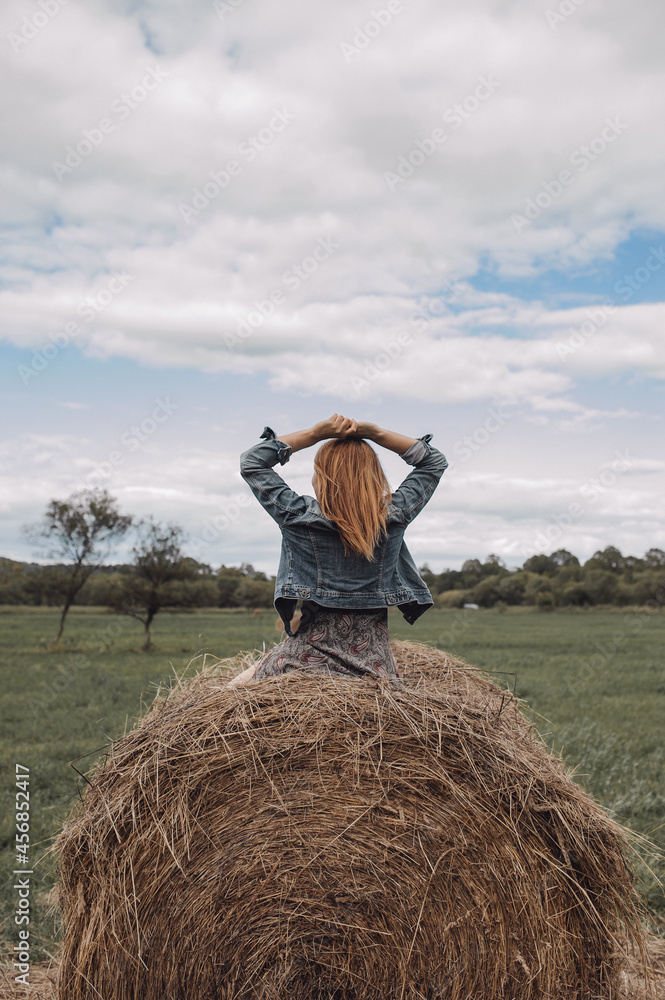 slender young woman is sitting on high haystack in field. rear view. red hair. freedom of travel. autumn landscape. selective focus slender young woman is sitting on high haystack in field. rear view. red hair. freedom of travel. autumn landscape. selective focus