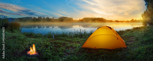 Orange tent with bonfire on bank of misty river at sunset