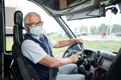 Portrait of senior white-haired bus driver in eyeglasses and facial mask fasten seat belt sitting at wheel