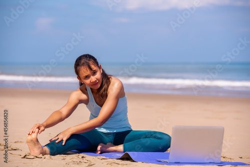 people, fitness, sport and healthy lifestyle concept - young asian woman watching in laptop and making yoga on tropical beach with blue sky background